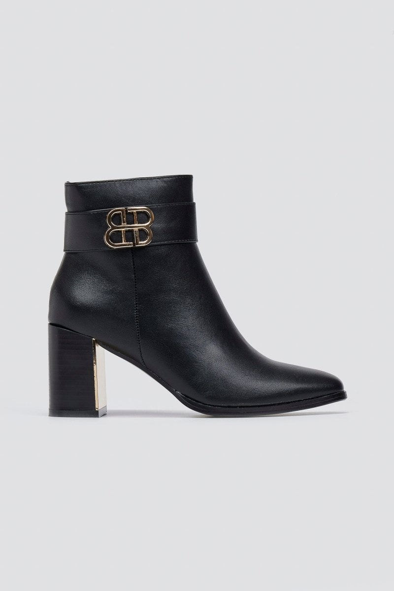 Black leather ankle boot with pointed toe, block heel and gold-tone logo buckle, angled view on a light grey background