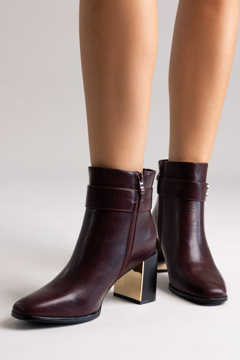 Burgundy leather ankle boots with pointed toe and gold-tone logo buckle, pair in front view on a model's legs in studio light