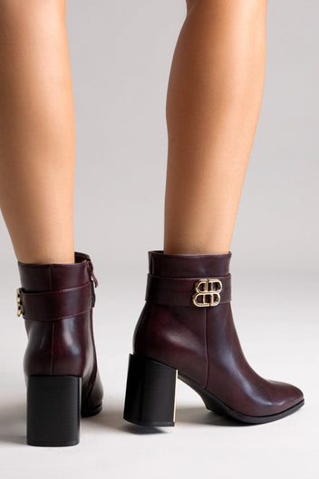Burgundy leather ankle boots with block heel and gold-tone logo buckle, pair in angled view on a model's legs in studio light