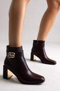 Burgundy leather ankle boots with block heel and gold-tone logo buckle, pair in front view on a model's legs in studio light