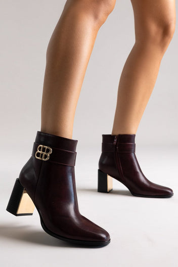 Burgundy leather ankle boots with block heel and gold-tone logo buckle, pair in angled view on a model's legs in studio light