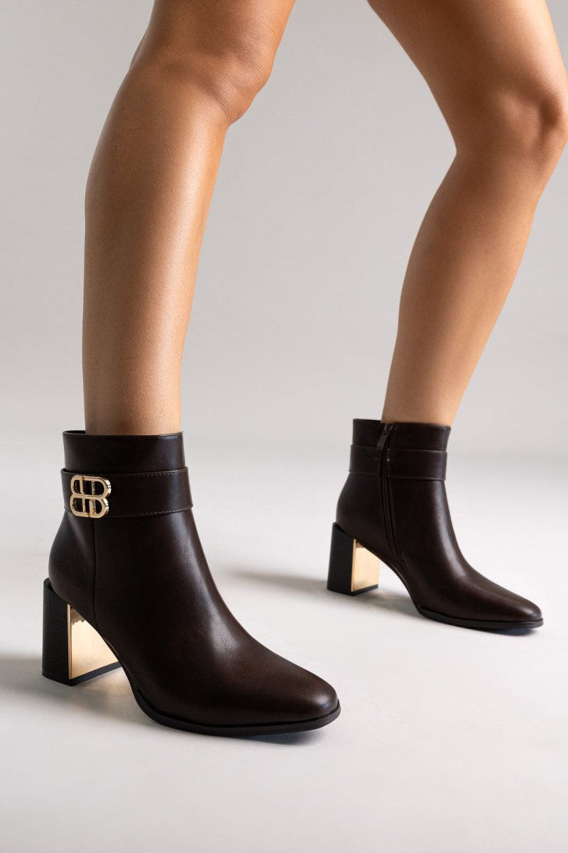 Dark brown leather ankle boots with pointed toe and gold-tone logo buckle, pair in angled view on a model's legs in studio light