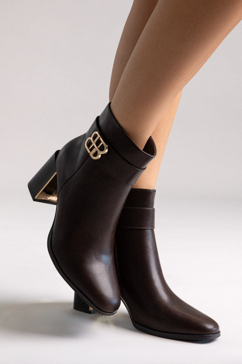 Dark brown leather ankle boot with block heel and gold-tone logo buckle, angled view on a model's legs in studio light