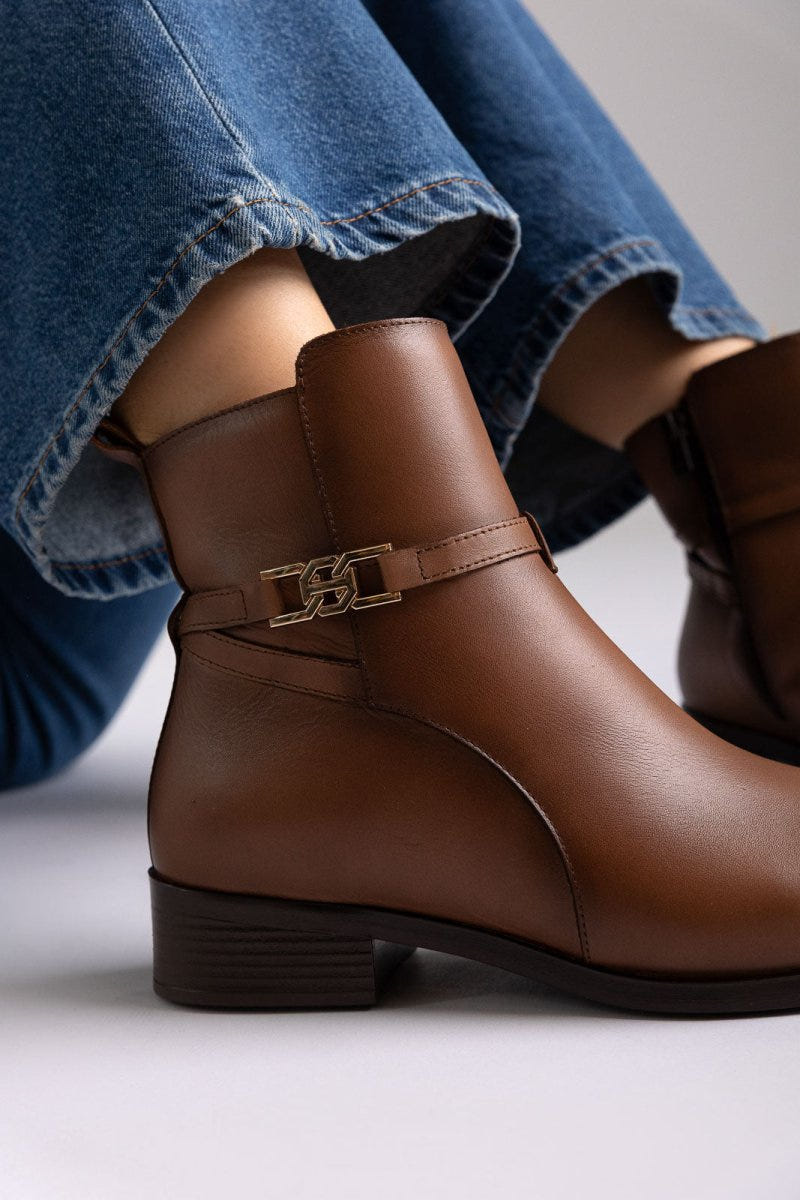 Brown leather ankle boot with low block heel and gold-tone buckle strap, angled view on a model's legs in studio light