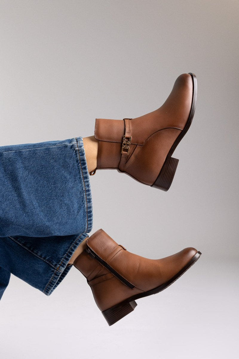 Brown leather ankle boots with flat heel and gold-tone buckle strap, pair in angled view on a model's legs in studio light