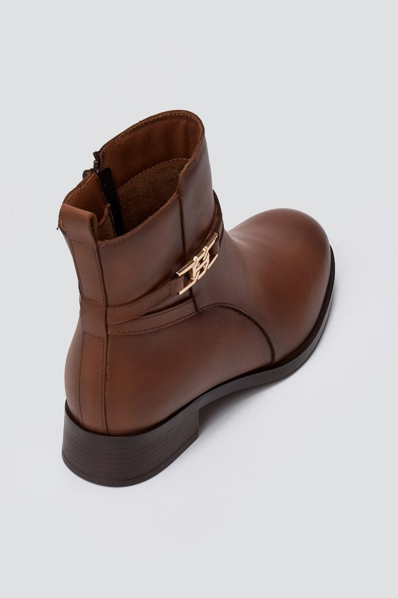 Brown leather ankle boot with flat heel and gold-tone logo buckle, angled view on a light grey background