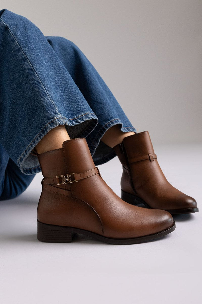 Brown leather ankle boot with low block heel and gold-tone buckle strap, pair in angled view on a model's legs in studio light