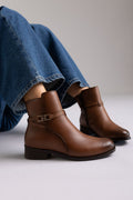 Brown leather ankle boot with low block heel and gold-tone buckle strap, pair in angled view on a model's legs in studio light