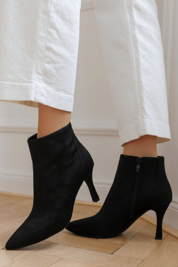 Black suede pointed-toe ankle boots with stiletto heel and side zip, pair in angled view on a model wearing white trousers