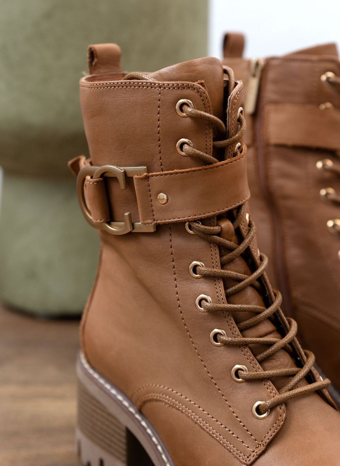 Close-up of tan leather lace-up boot with metal buckle straps and lug sole, detail view on a light background
