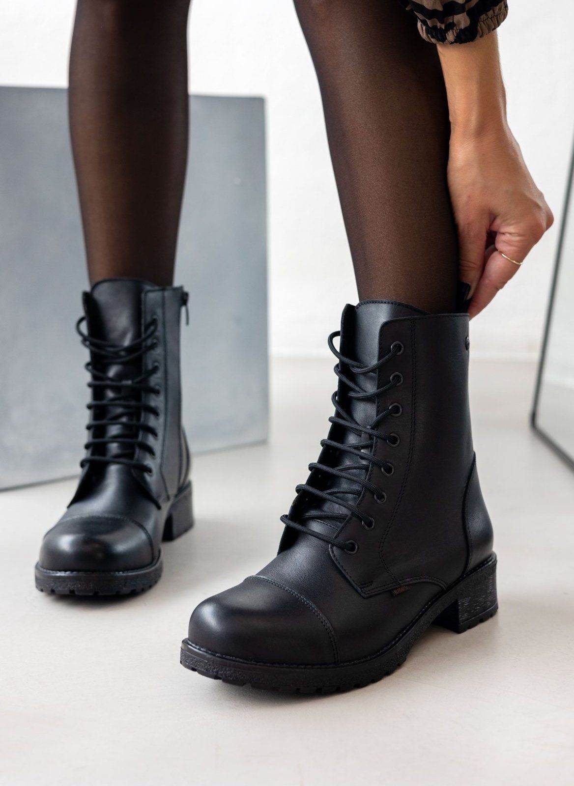 Black leather lace-up ankle boots with lug sole, pair in front view on a model's legs and sheer black tights in studio light