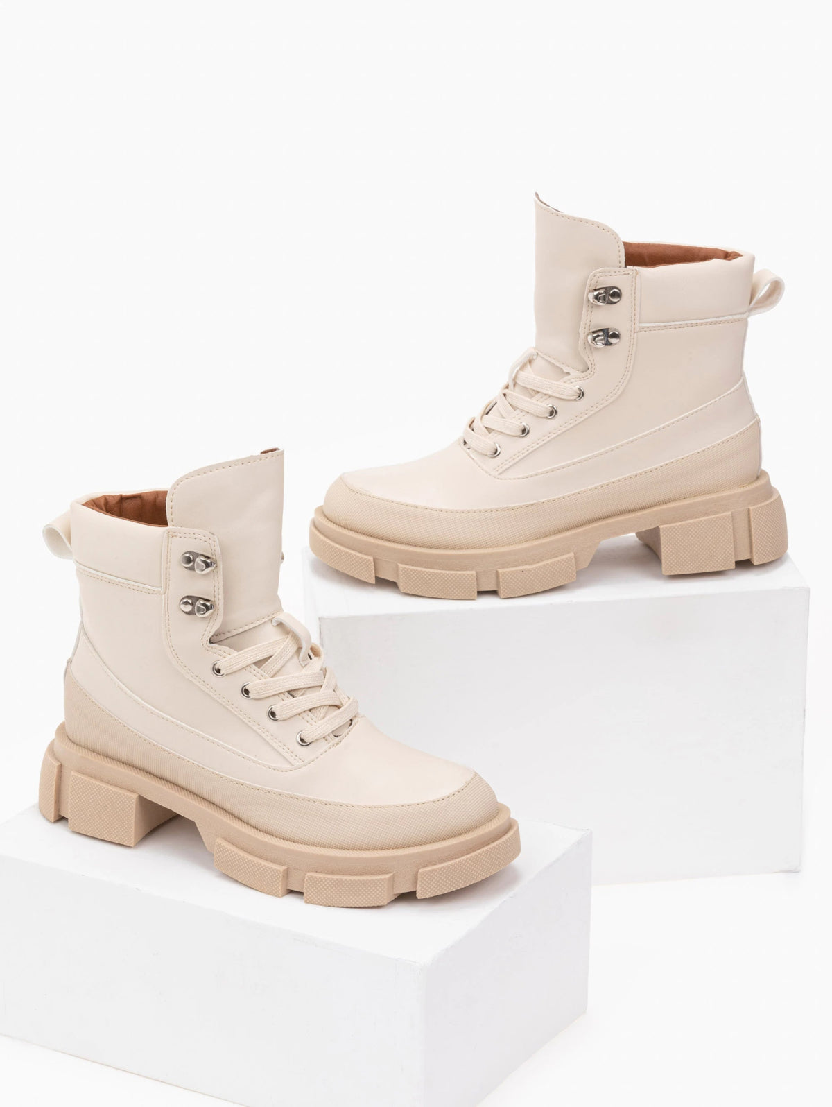 Pair of beige faux leather lace-up ankle boots with metal eyelets, tan lining and chunky lugged soles, displayed on white blocks