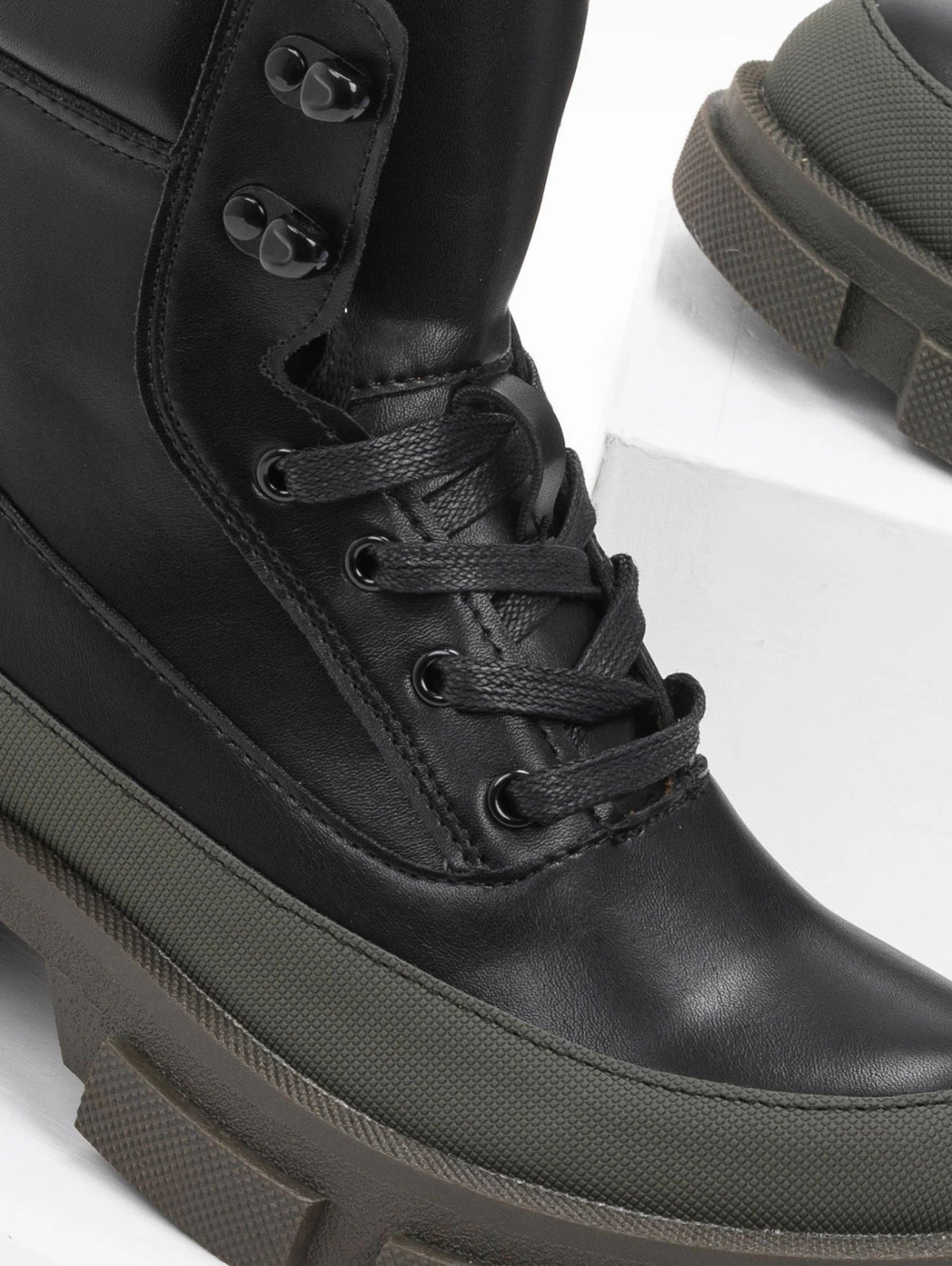 Close-up of black faux leather lace-up ankle boot with black laces, metal eyelets, khaki green toe panel and lugged sole