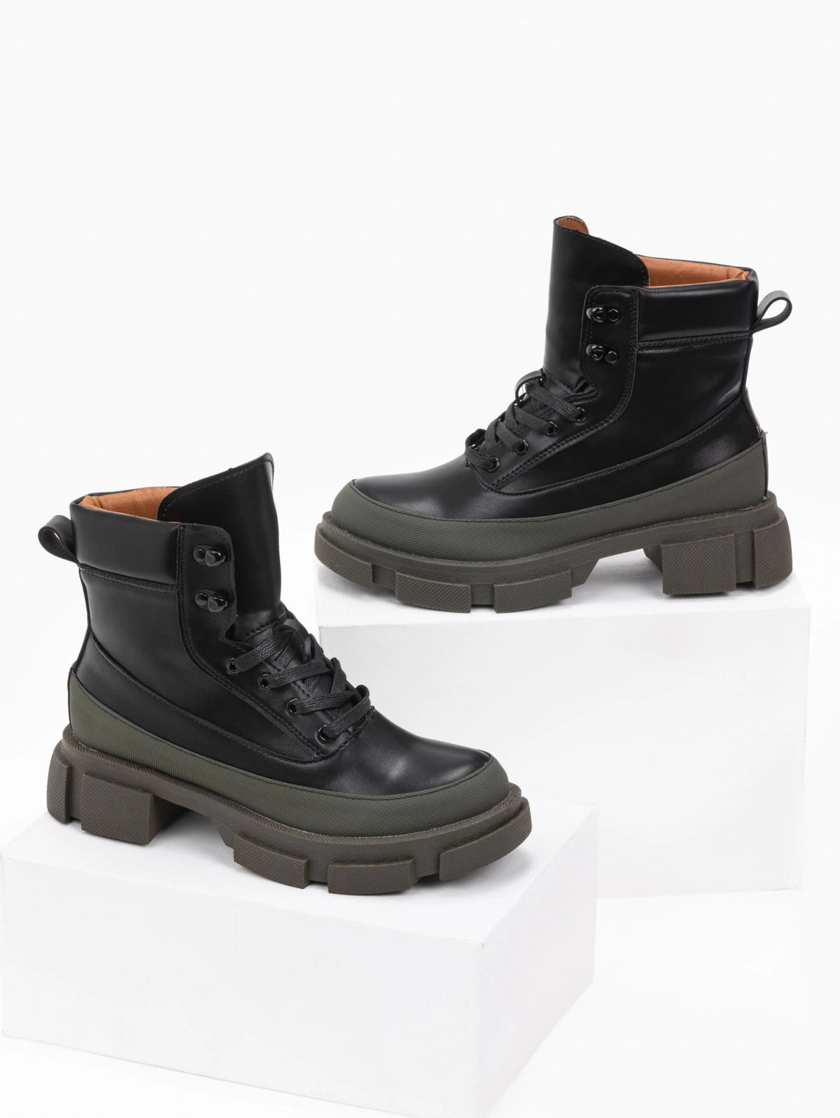 Pair of black faux leather lace-up ankle boots with khaki green toe caps, tan lining and chunky lugged soles, displayed on white blocks