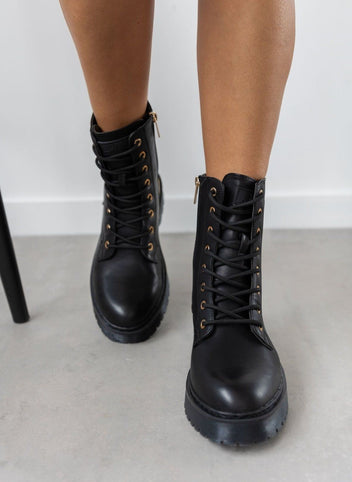 Model wearing black lace-up boots with chunky sole and gold eyelets, front view of feet on a light grey floor