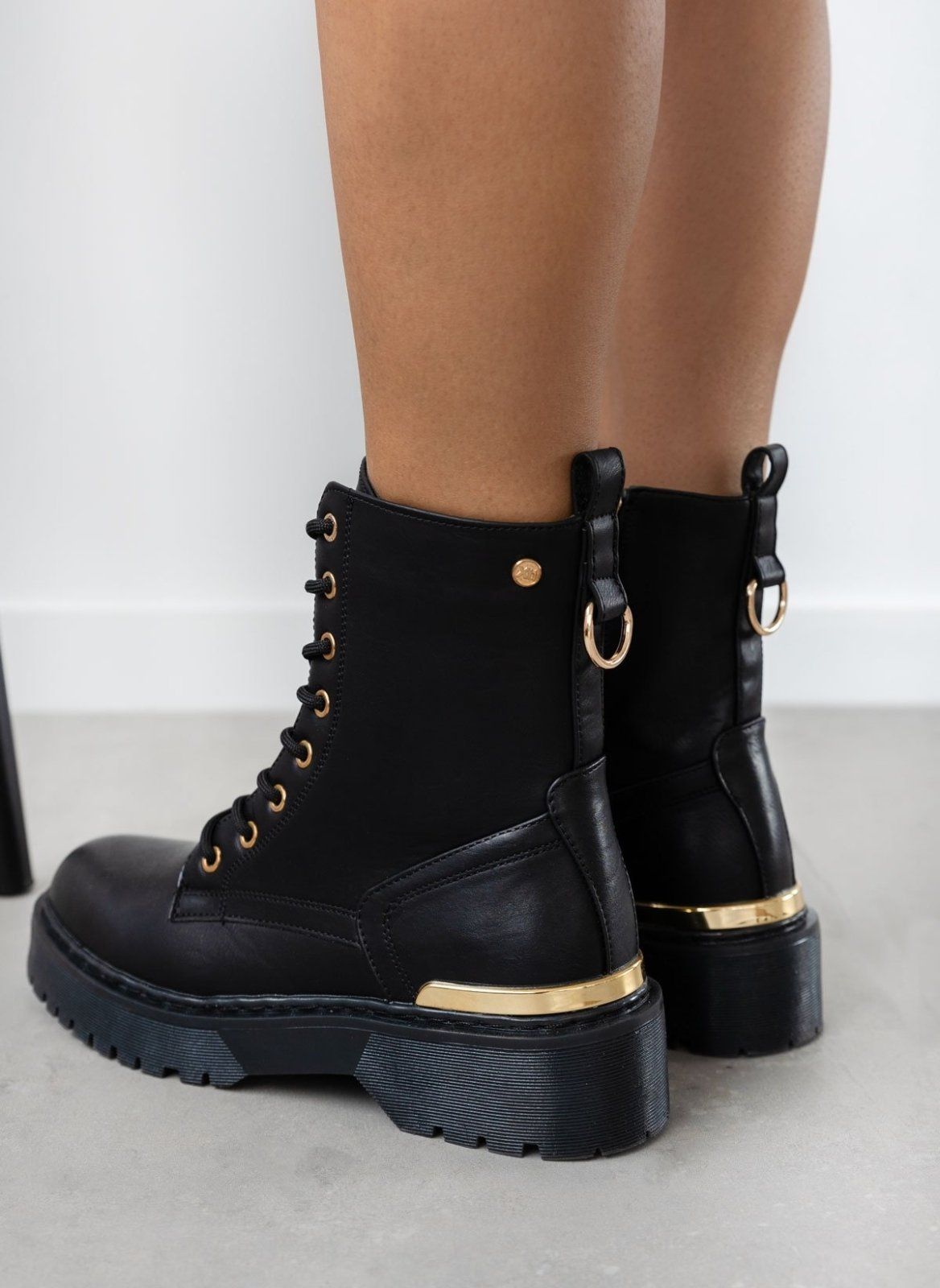 Model wearing black lace-up boots with gold eyelets, gold trim and chunky sole, front three-quarter view