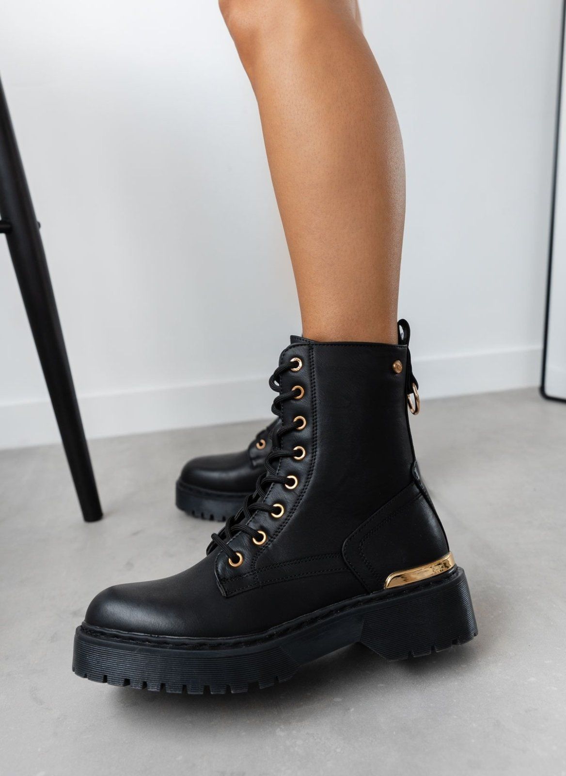 Model wearing black lace-up boot with gold eyelets and chunky sole, side view on grey floor