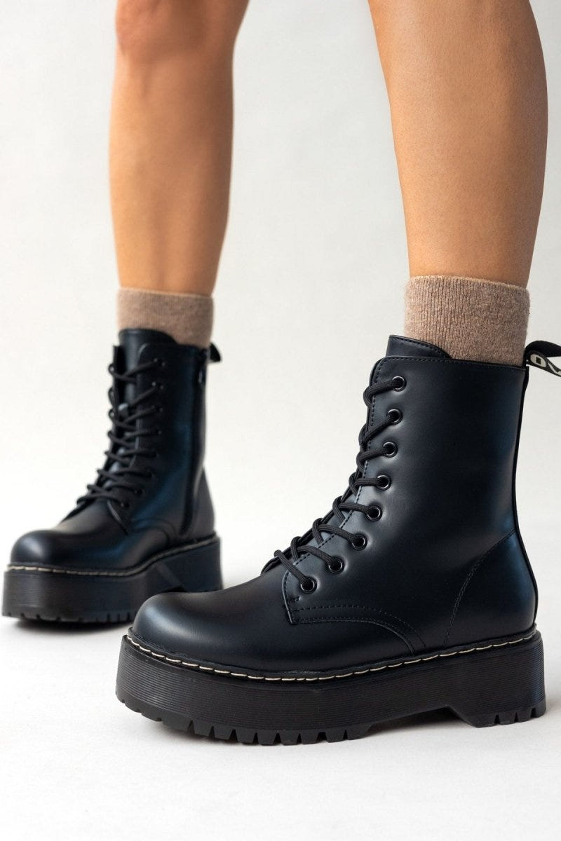 Model wearing black leather lace-up boots with chunky sole and white stitching over beige socks