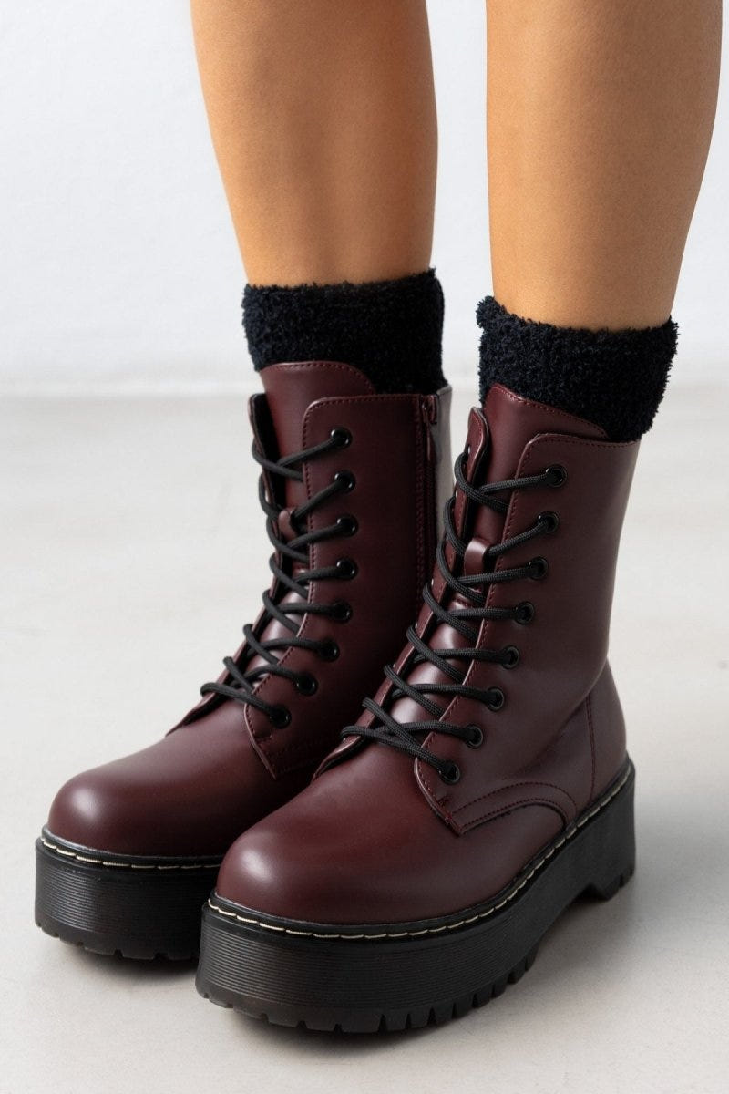 Model wearing burgundy leather lace-up boots with chunky black sole over black knit socks
