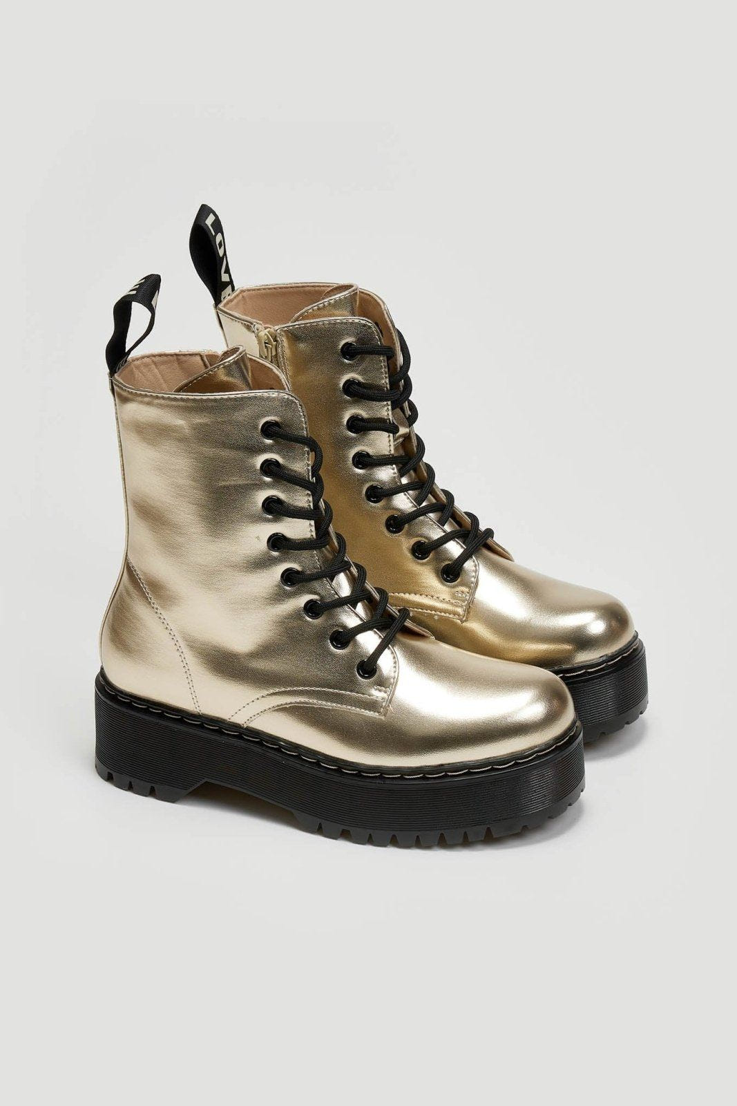 Pair of gold metallic lace-up boots with black laces and chunky black sole on a light grey background