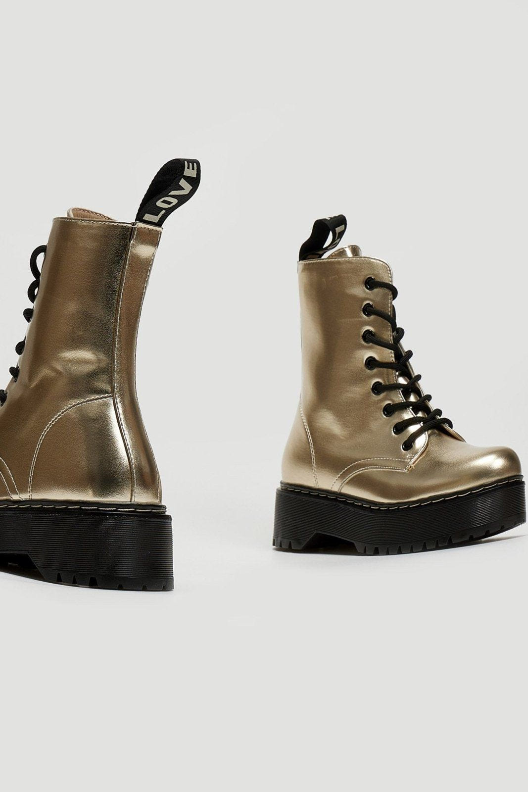 Pair of gold metallic lace-up boots with black laces and chunky black sole on a light grey background