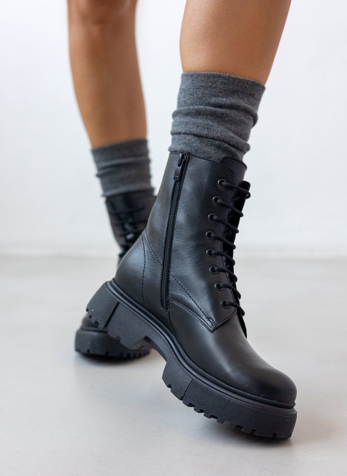 Black leather lace-up ankle boots with lug sole and side zip, pair in angled view on a model's legs and grey socks in studio light