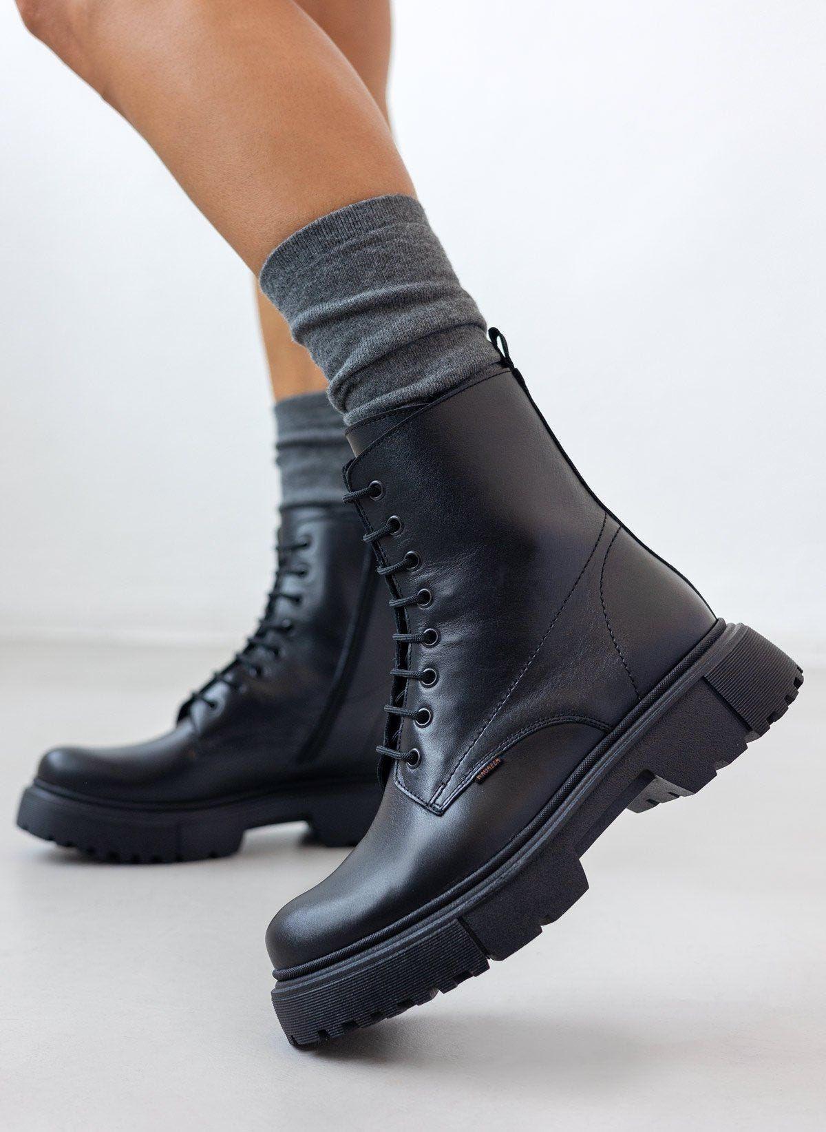 Black leather lace-up ankle boots with lug sole and side zip, pair in front view on a model's legs and grey socks outdoors