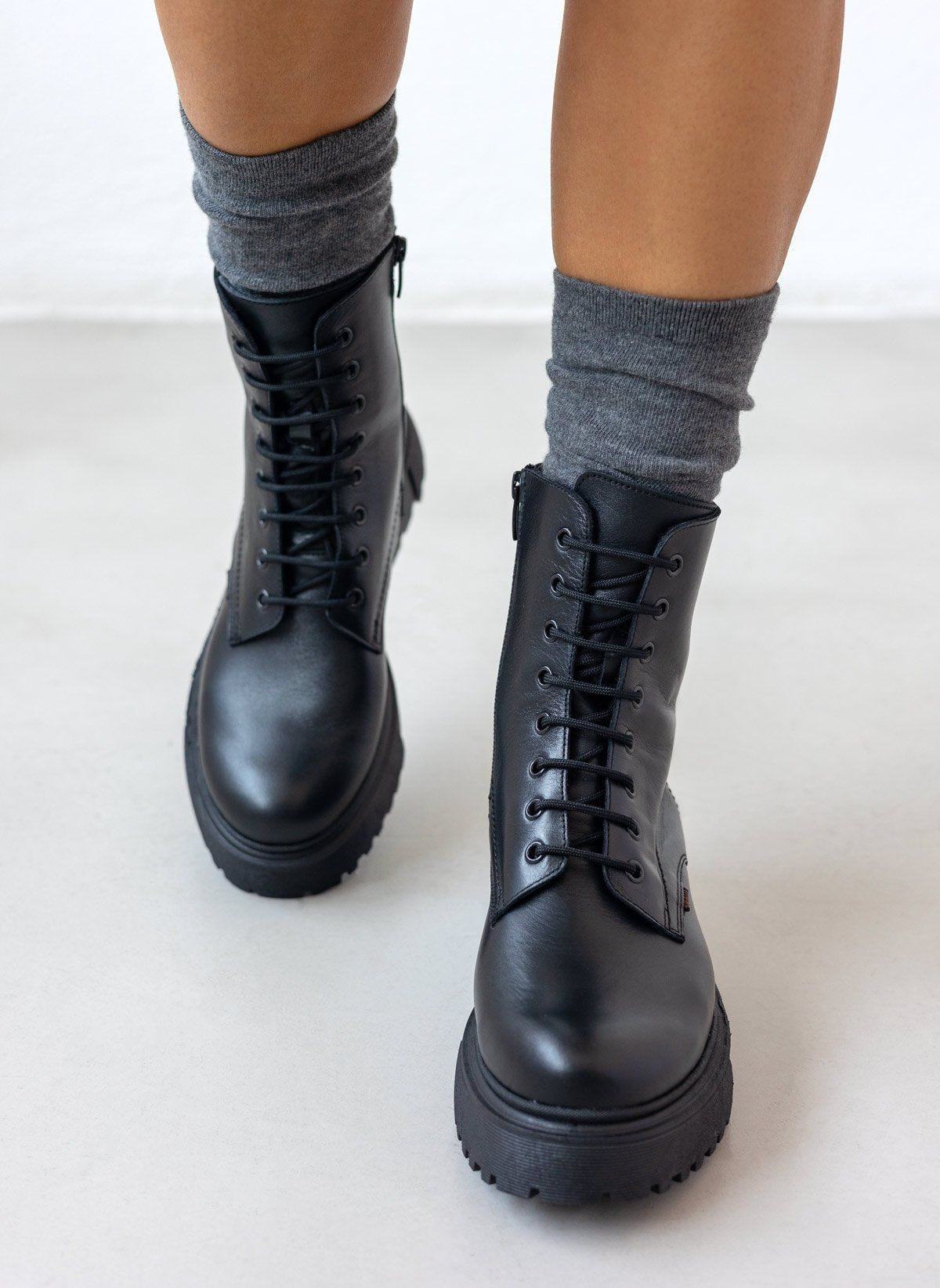 Black leather lace-up ankle boots with lug sole, pair in angled view on a model's legs and grey socks in studio light