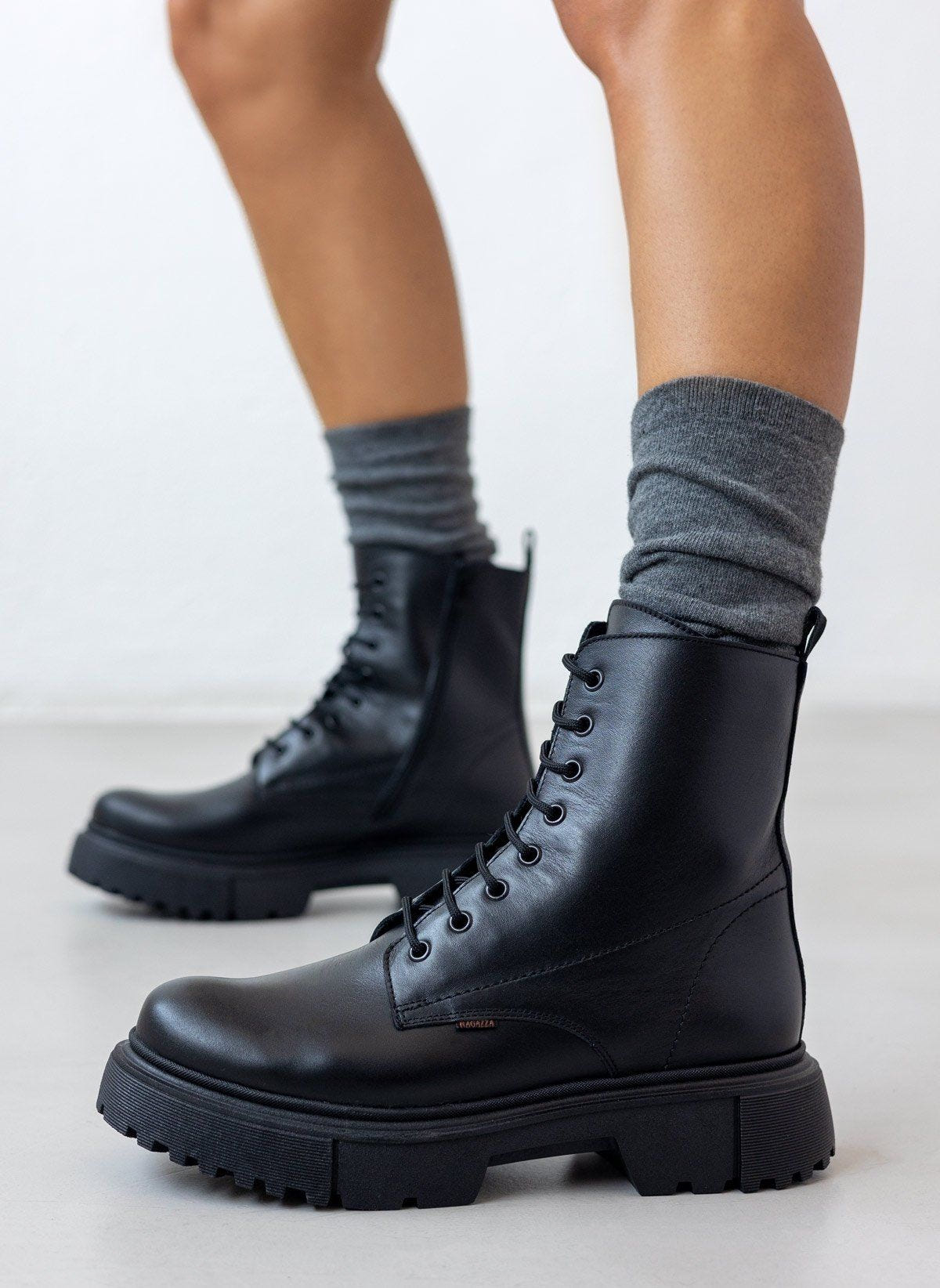 Black leather lace-up ankle boots with lug sole, pair in side view on a model's legs and grey socks in studio light