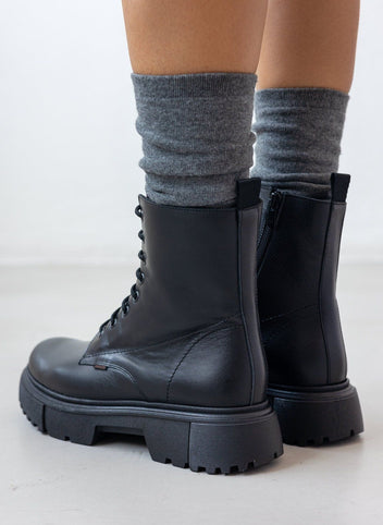 Black leather lace-up ankle boots with lug sole, pair in front view on a model's legs and grey socks in studio light