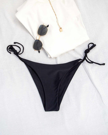 Black side-tie bikini bottom with white top, gold necklace and sunglasses laid flat on white background