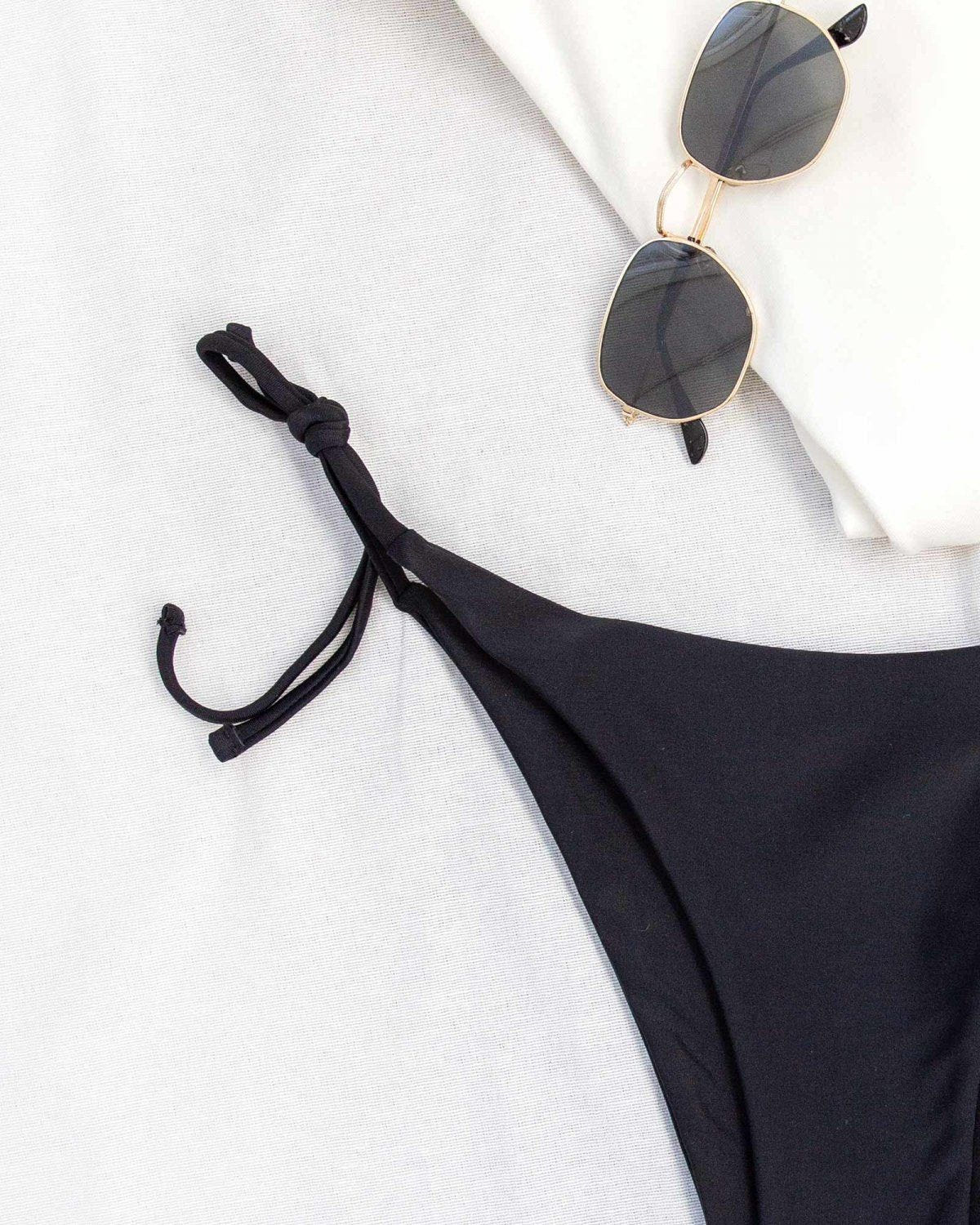 Close-up of black side-tie bikini bottom laid flat on white with sunglasses and a gold chain