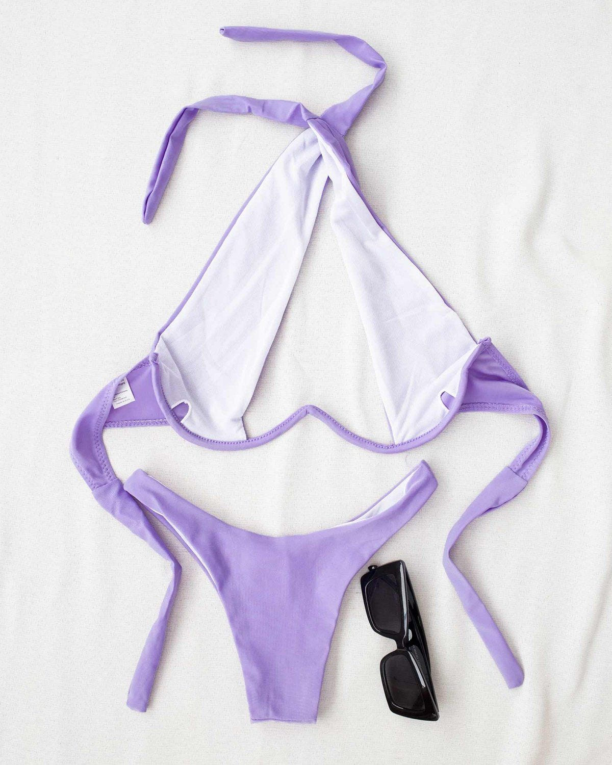 Lilac halter bikini with cutout detail and tie straps laid flat on a white background beside sunglasses
