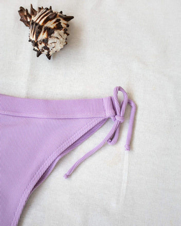 Lilac side-tie bikini bottom with tie sides, laid flat on white background beside a seashell