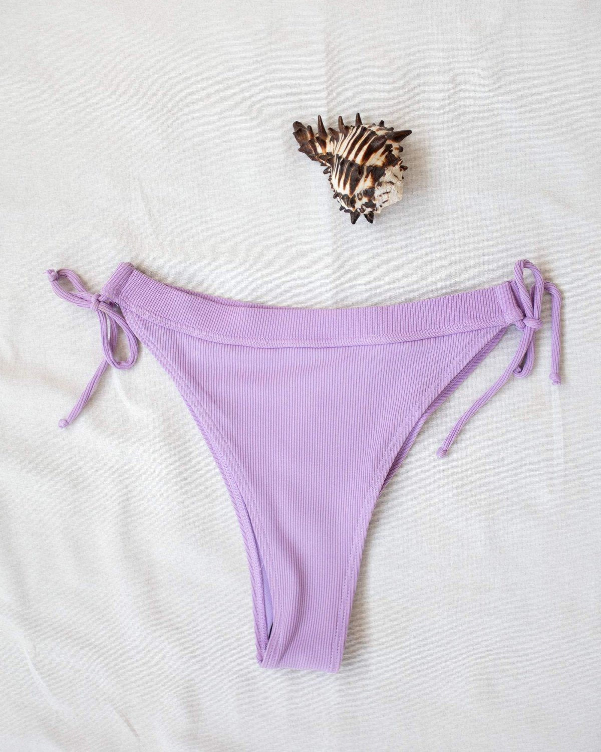 Lilac side-tie bikini bottom with tie sides, laid flat on white background beside a seashell