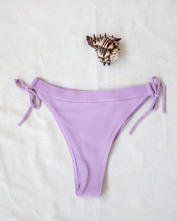 Lilac side-tie bikini bottom with tie sides, laid flat on white background beside a seashell