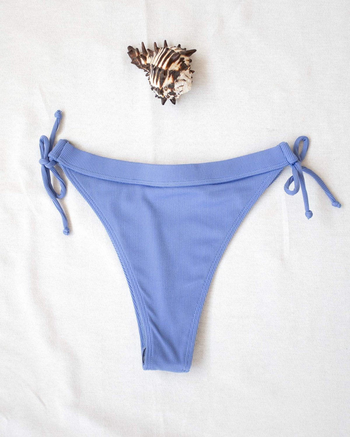 Periwinkle blue ribbed side-tie bikini bottom with tie straps, laid flat on white background beside a seashell
