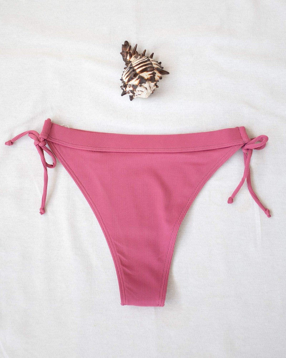 Pink ribbed side-tie bikini bottom with tie straps, laid flat on white background beside a seashell