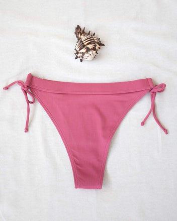 Pink ribbed side-tie bikini bottom with tie straps, laid flat on white background beside a seashell