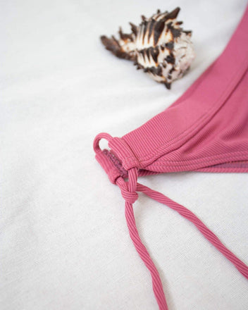 Pink ribbed side-tie bikini bottom with tie straps, laid flat on white background beside a seashell