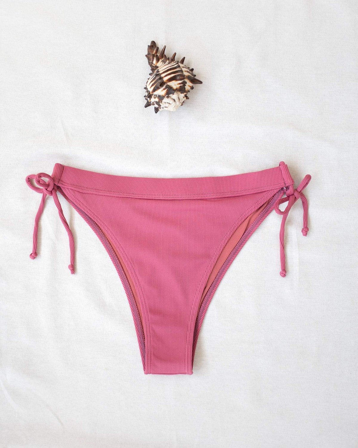 Pink ribbed side-tie bikini bottom with tie straps, laid flat on white background beside a seashell
