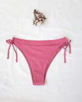 Pink ribbed side-tie bikini bottom with tie straps, laid flat on white background beside a seashell