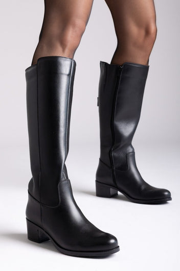 Black leather knee-high boots with side zip and block heel, close-up of legs on light backdrop, angled view