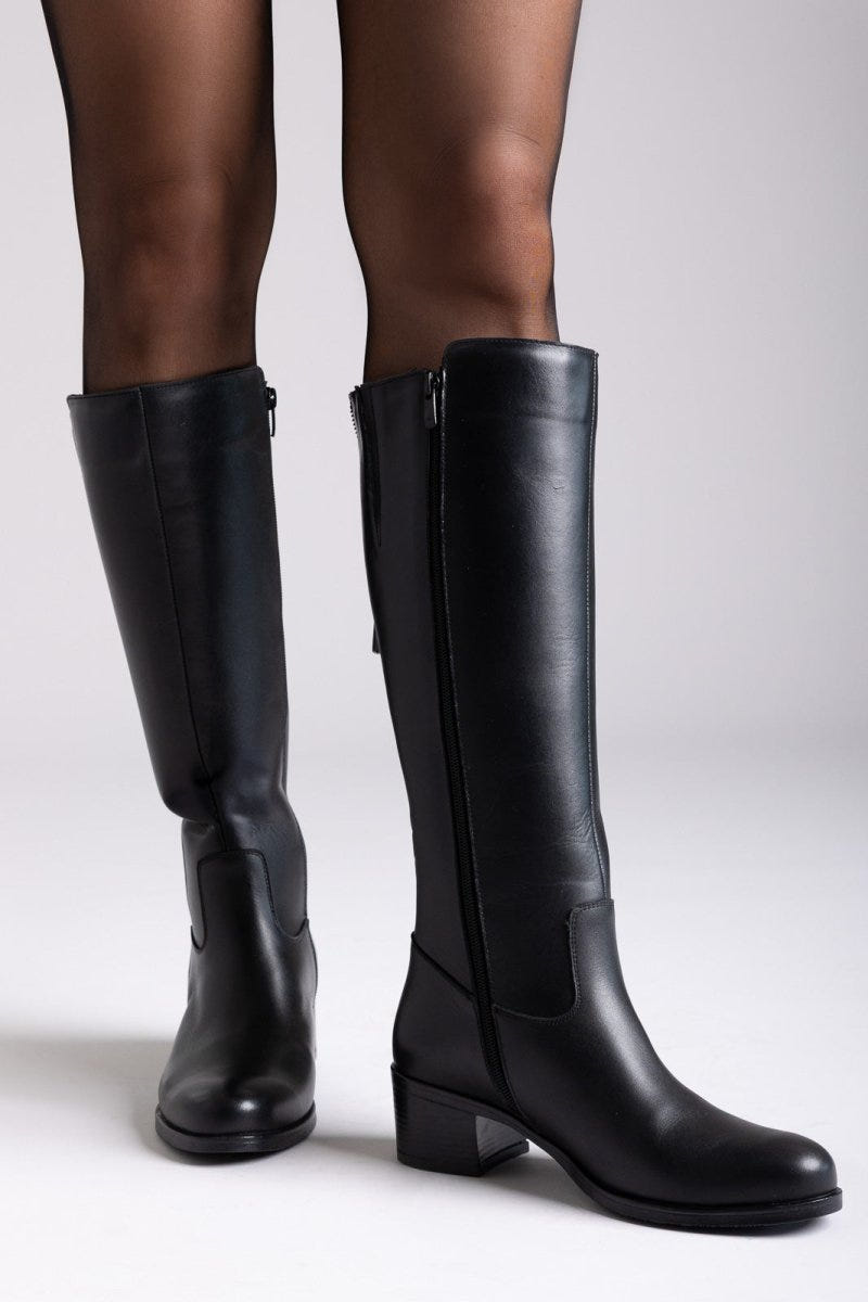 Black leather knee-high boots with side zip and block heel, close-up of legs on light backdrop