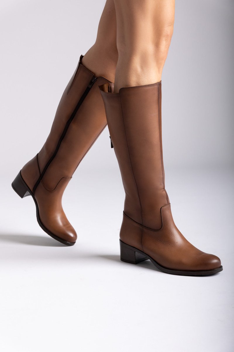Tan leather knee-high boots with side zip and block heel, close-up of legs on grey backdrop