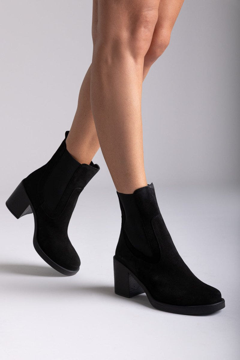 Model legs wearing black suede ankle boots with block heel and side panels, close-up on grey backdrop