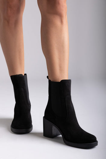 Model legs wearing black suede ankle boots with block heel and side panels, close-up on grey backdrop, front view