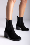 Model legs wearing black suede ankle boots with block heel and side panels, close-up on grey backdrop, side view