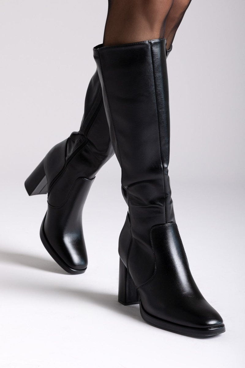 Model legs in black knee-high block heel boots with smooth leather shafts, worn with dark tights, angled view on a light grey backdrop