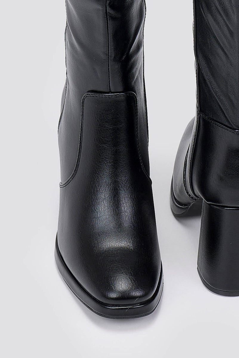 Close-up of black knee-high boot with a block heel and square toe, showing the shaft and heel on a light grey backdrop